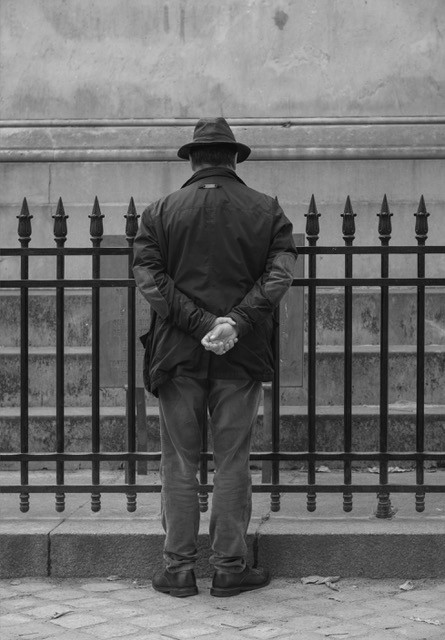 Black and white photograph — man in a hat standing at an iron fence